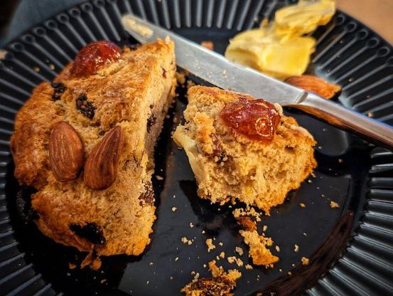 A photo of a golden baked cake thing, known as a Fat Rascal, cut in half on a blue plate, served with butter. A knife is cunningly positioned for further butter application. The Fat Rascal has cherries for eyes and almonds for teeth.