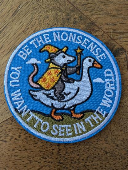 in my wooden table my new sticker embroidery showing a white goose ridden by a mystic wearing a cape and a wand, surrounded by the words BE THE NONSENSE YOU WANT TO SEEN IN THE WORLD