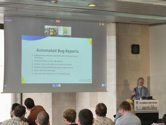John Harrison stands at a lectern in the lower right corner of the photo, looking a bit downward as he gestures. On a projection screen to John's right is a slide from his talk, titled "Automated Bug Reports" and then a series of steps:

1. Automatically generate a dump file with all relevant kernel information
2. Send notification to user side daemon
3. Daemon collects all useful non-kernel information
4. Daemon sends collected bug report to global server
5. Server does first step triage