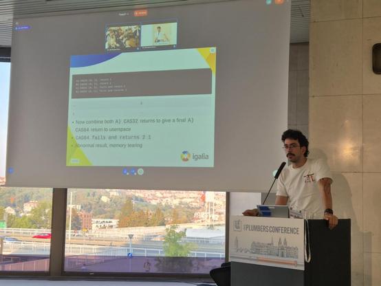 André Almeida presenting "Emulating x86 atomics on arm64". André is standing at a lectern to the right of the image, looking out into the audience. To his right and our left, there is a projection screen showing a slide from his talk.