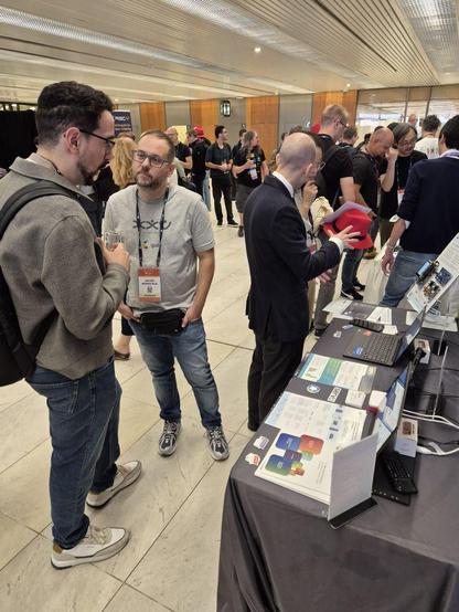 A small crowd of people standing in front of a table and having a number of conversations.Beyond them are still more people standing and talking or walking past.