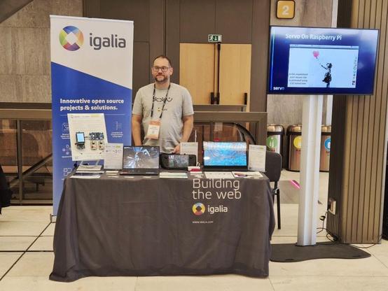 Igalia’s booth at OSS EU, which is a table covered with stickers to take, various bits of printed material, and a laptop. Behind it stands an Igalian, and a monitor on a stand showing a video about Servo on Raspberry Pi.