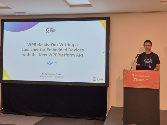 Mario stands smiling at a lectern, next to a projection screen bearing the title slide from Mario’s talk, “WPE Hands-On: Writing a Launcher for Embedded Devices with the New WPEPlatform API”.