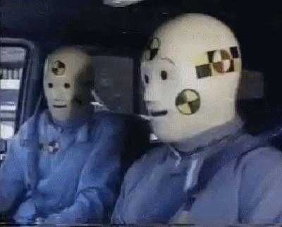 The image depicts two crash test dummies seated inside a vehicle, both wearing light blue clothing and matching rubber masks with safety markings.
