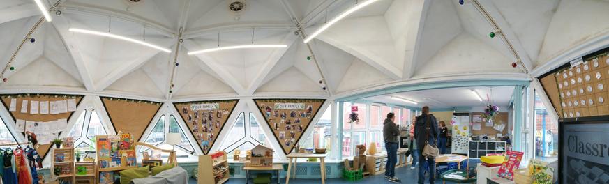 A panoramic photo of a primary school classroom. The upper half shows hollow triangular pyramids with four strip lights and one fan visible. The lower half shows triangular windows and triangular noticeboards decorated with photos and learning materials. Small classroom furniture is scattered around the edge of the room. Four adults are talking and standing to the right of middle at the entrance to the classroom (which is a more conventional rectangular area).