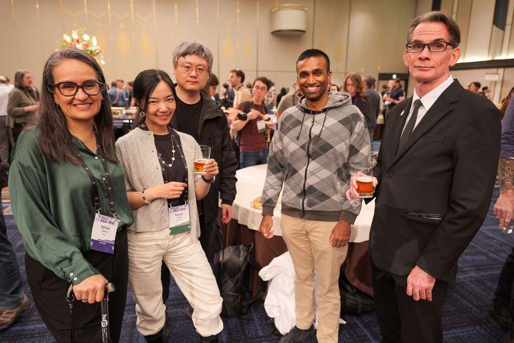 A group of 5 persons socializing at a reception; two women and three men. They wear casual and smart attire. All are smiling. Some hold drinks, In the background we see many other people who appear to be networking.