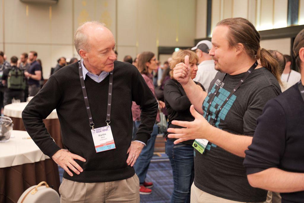 Two men engaged in conversation at a gathering. One is speaking and the other is listening while looking at his name tag. Several people are visible in the background, suggesting a networking event.