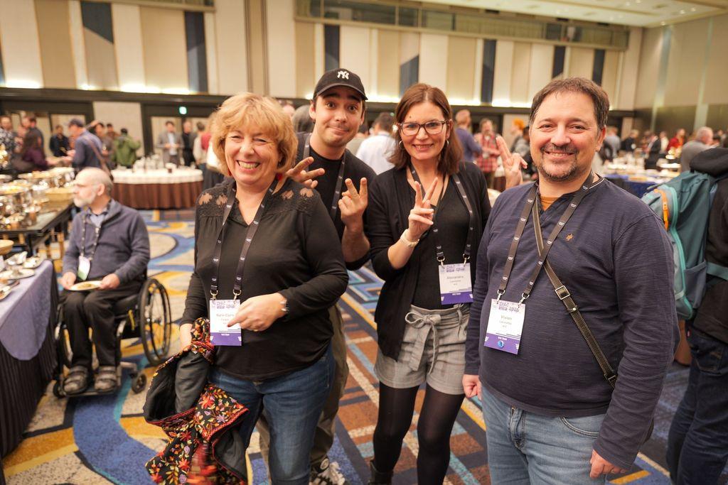 A group of four people posing at an event. They are all smiling and some are making peace signs. The background is crowded with other people who are at the event. A person in a wheelchair is visible right behind the group next to a food table.