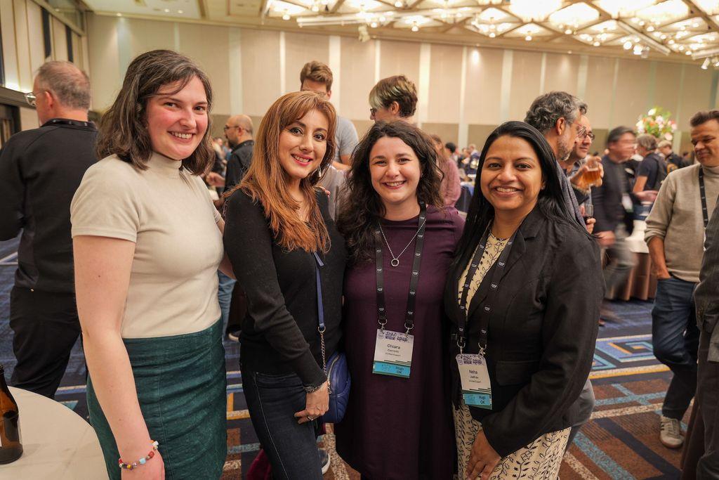 Four women stand together smiling, in a lively social event setting. They are dressed in a mix of casual and professional attire with name badges visible. In the background, other guests are engaged in conversation.